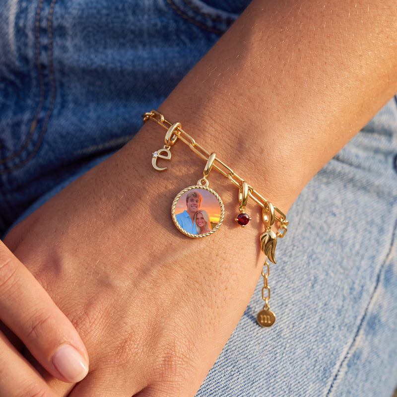 Wrist resting on faded denim, wearing a gold paper clip charm bracelet with an initial e, a round photo charm of a young couple at sunset, a garnet birthstone and an angel wing
