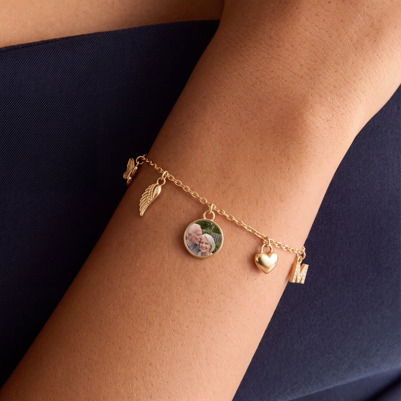 Wrist wearing a dainty gold mini charm bracelet with a butterfly, an angel wing, a round photo charm of an older couple, a puffy heart and a pave M