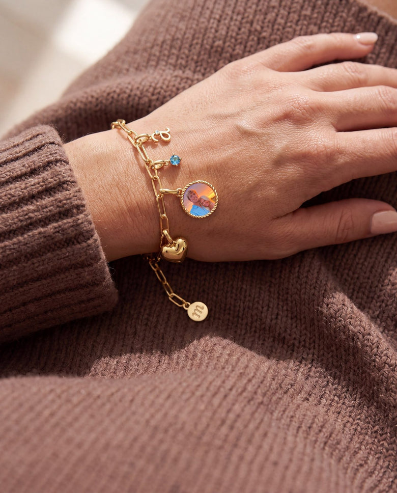 Hand on a brown sweater wearing a gold paperclip charm bracelet with an initial, a birthstone, a heart and a couple's photo charm
